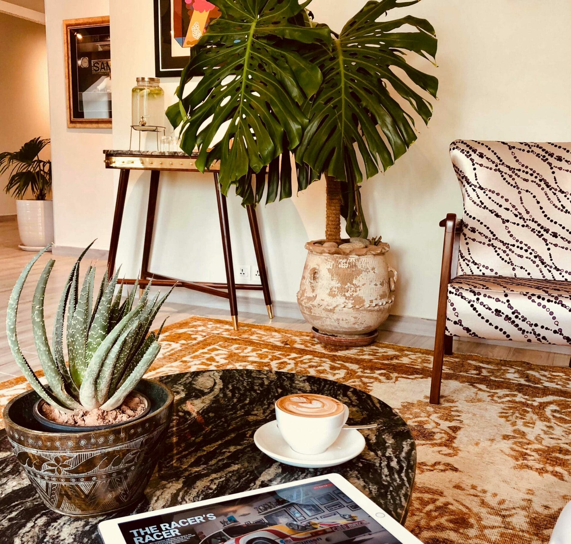 A stylish landing features plants, a cup of coffee, and a tablet on a marble table. An upholstered chair and a wooden side table with a glass container are in the background. - The 108 Hotel Islamabad