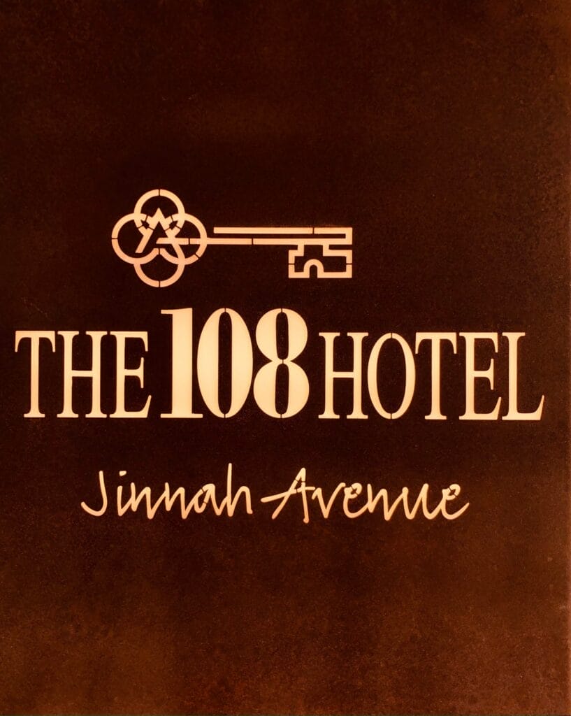 Sign reads "The 108 Hotel Jinnah Avenue" with a key symbol above the text. The background, a deep dark brown, evokes the elegance of an art gallery. - The 108 Hotel Islamabad