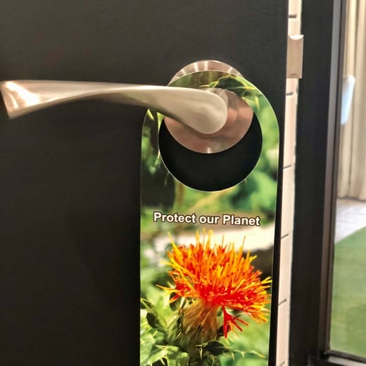 A green door hanger with a photo of a bright orange flower hangs from a silver door handle. The text on the hanger reads "Protect our Planet," reminding us that small actions in our rooms can make a big difference. - The 108 Hotel Islamabad