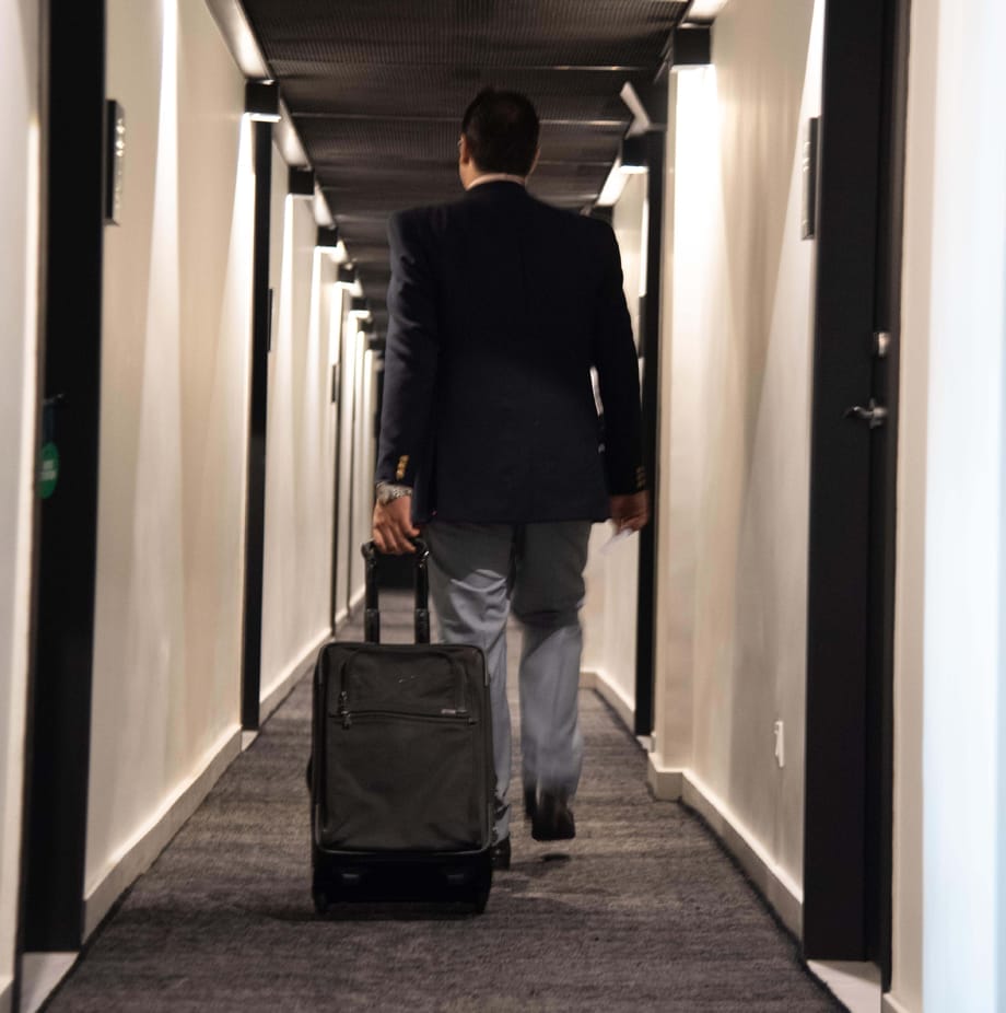 A man in a suit walks down a narrow hallway with a rolling suitcase, likely heading towards his accommodations. - The 108 Hotel Islamabad