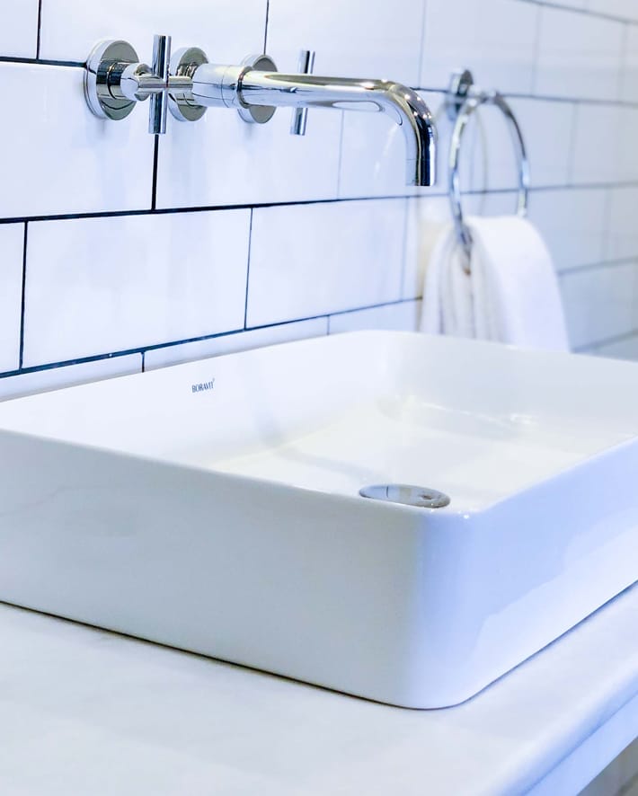 A modern bathroom sink with a square white basin and a chrome wall-mounted faucet. In this well-designed room, a white towel hangs on a round chrome towel ring attached to the tiled wall. - The 108 Hotel Islamabad