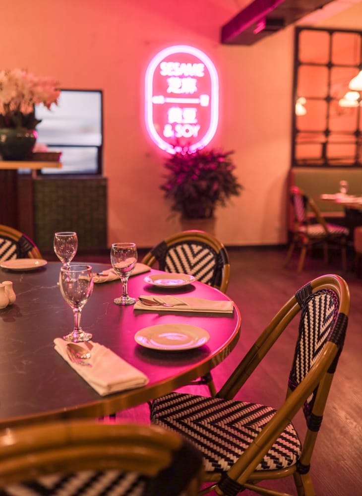 A dimly lit restaurant scene with a round table set for dining, featuring neatly arranged napkins, wine glasses, and plates. A neon sign in the background reads "Sesame," while patrons enjoy their food and drink at "Rice & Soy. - The 108 Hotel Islamabad