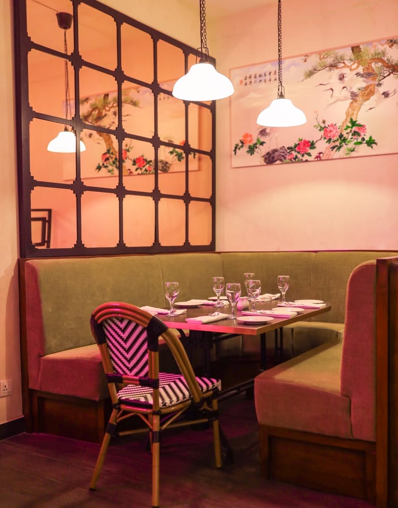 A restaurant booth with green cushions, a wooden table set with glassware ready for your favorite drink, and a pink-patterned chair. The area is lit by hanging lamps and decorated with a painting and a grid-style mirror. - The 108 Hotel Islamabad