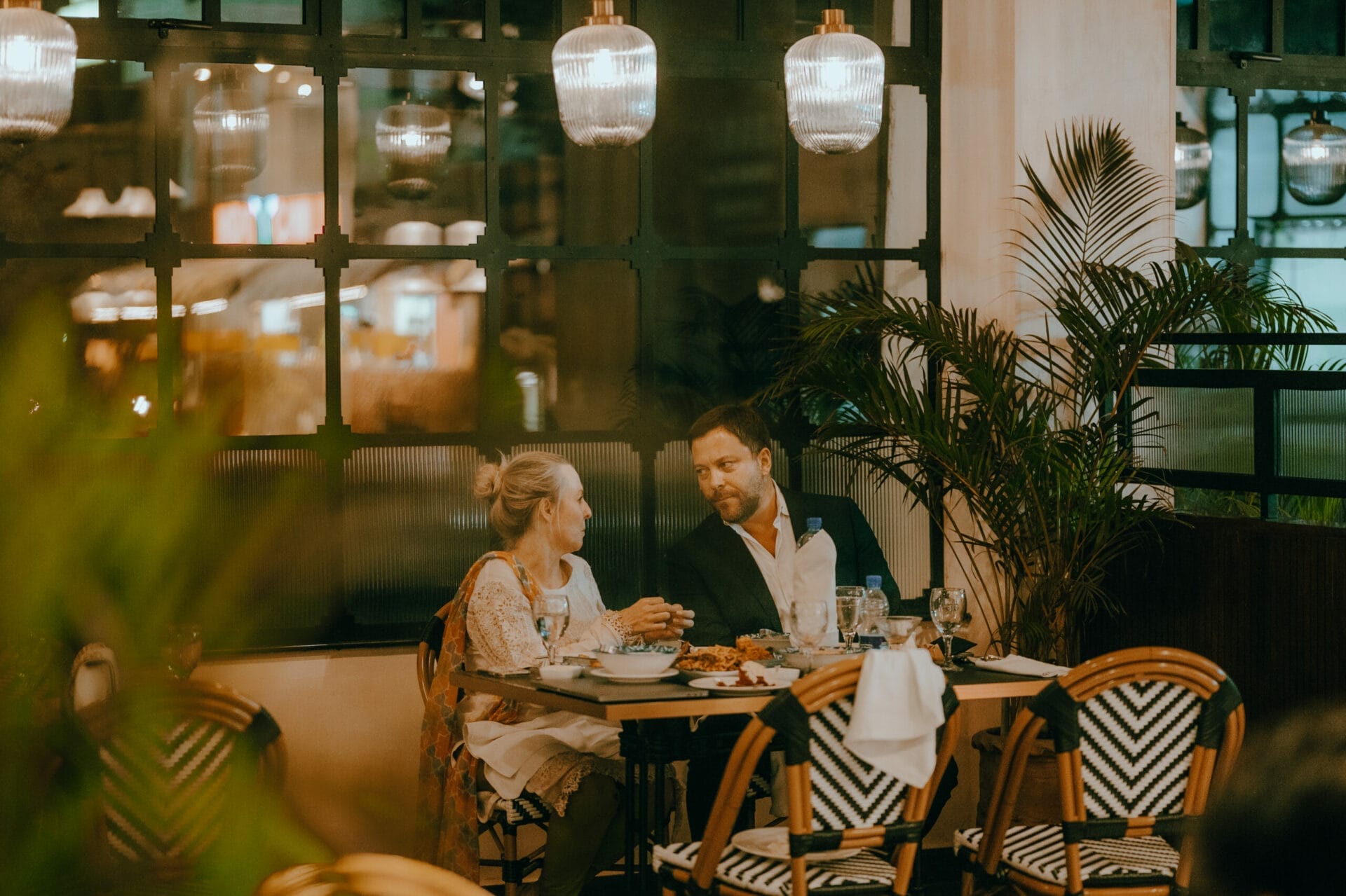 A man and a woman are sitting at a dining table in a restaurant, engaging in conversation. The table is set with delicious food and drinks. Decorative plants and pendant lights create a cozy atmosphere. - The 108 Hotel Islamabad