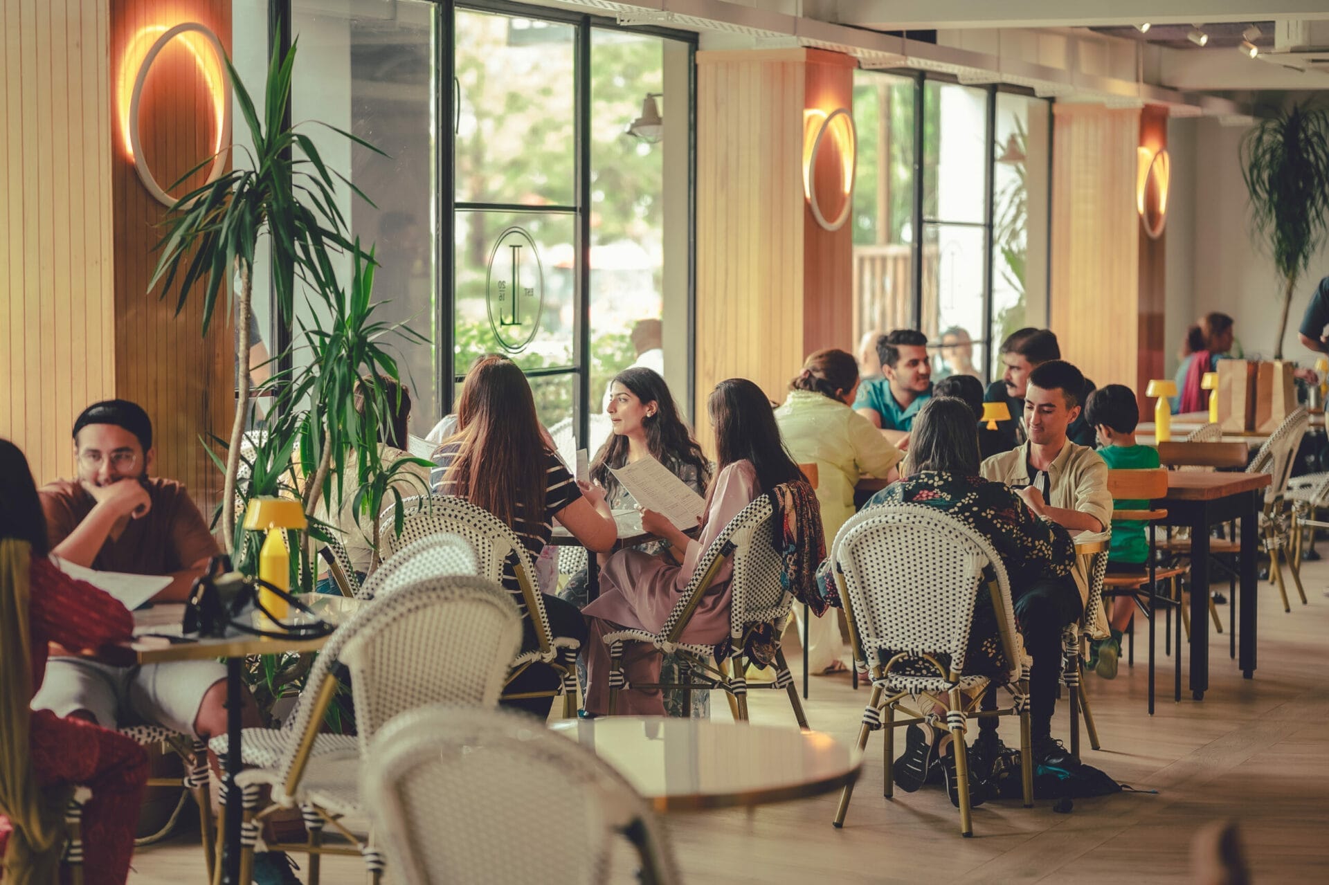 People seated at tables inside a café, talking and enjoying their meals and drinks. The café features large windows and warm lighting with decorative plants. - The 108 Hotel Islamabad