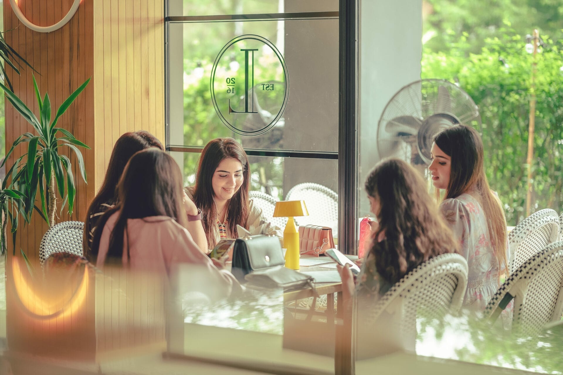 A group of women sit around a table in a cafe, with drinks and food neatly arranged alongside their electronic devices, engaged in animated conversation. - The 108 Hotel Islamabad