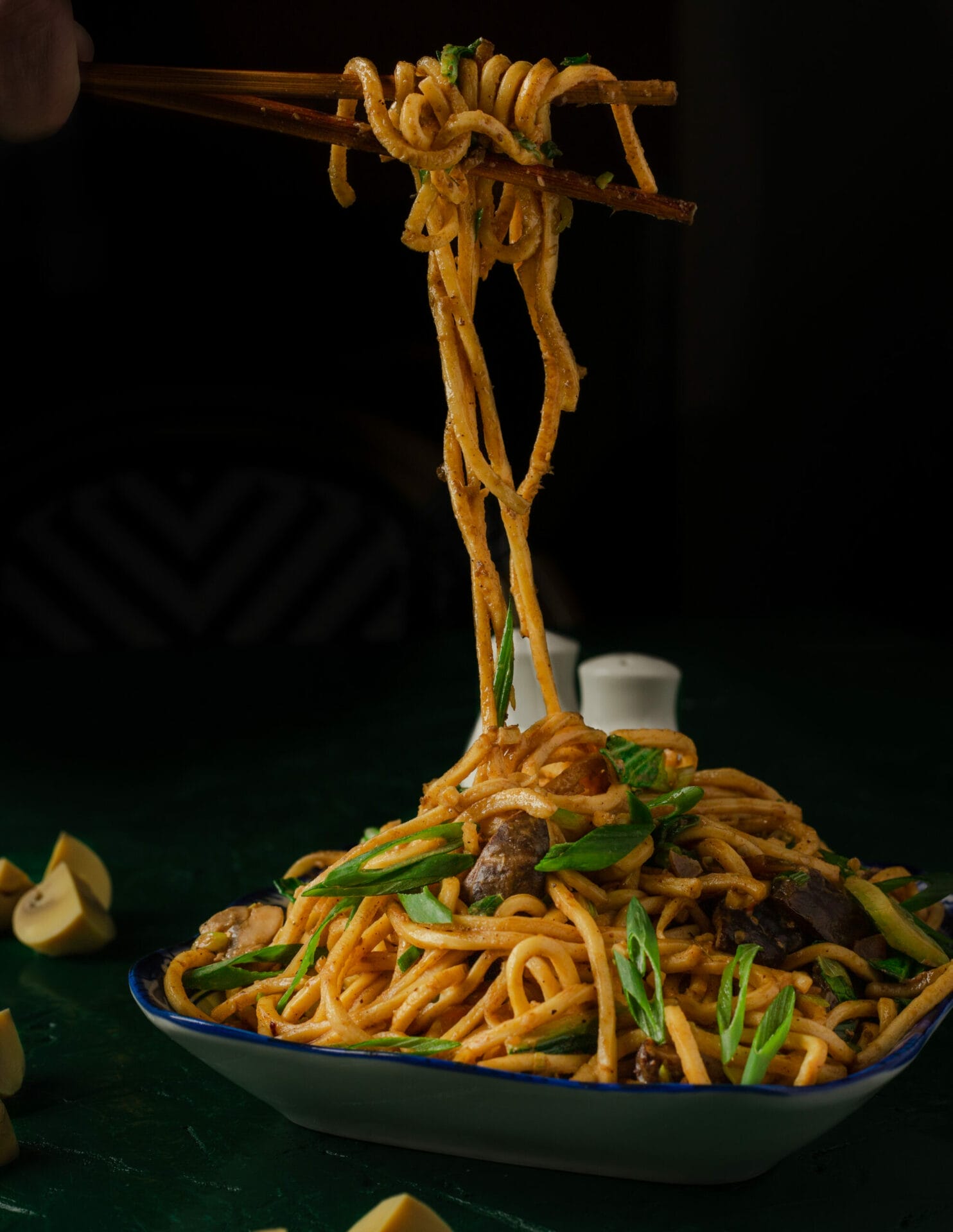 A pair of chopsticks holds up a portion of noodles from a bowl filled with delicious food, including vegetables and pieces of meat. The scene is set against a dark background. - The 108 Hotel Islamabad